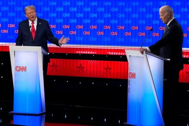 A man in a suit on the left stands behind a podium, that is light in a blue-white gradient, motio<em></em>ning with his left hand toward another man in a suit standing behind a similar podium.