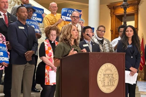 A woman is showing speaking at a podium from a distance, with a couple dozen people, both men and women, standing behind her. Some of those people hold placards whose words aren't entirely visible.