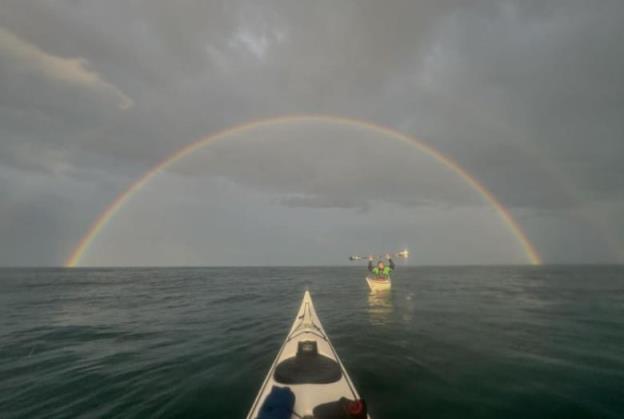 A bow of a kayak points toward a rainbow on the horizon. A man in another kayak holds his paddle above his head.