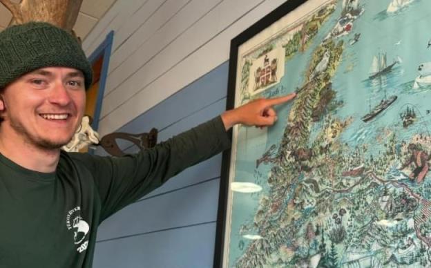 A man in a green shirt and hat points to map of Newfoundland mounted on a wall.