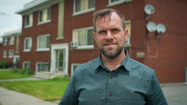 A man in a button-up shirt with a beard stands in front of a red-brick building