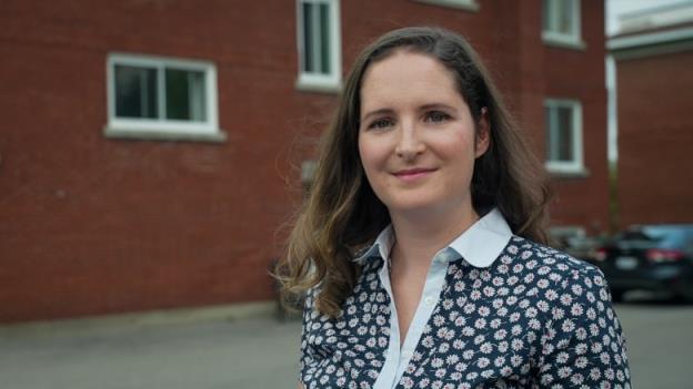 A woman in a blue and white floral shirt stands behind a red brick building. 