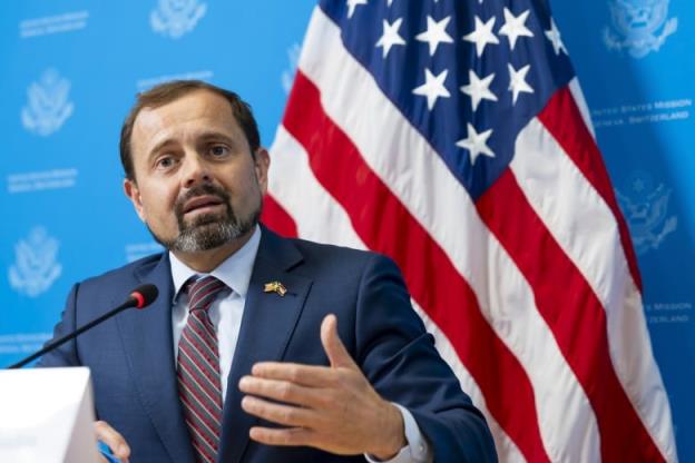 A bearded man wearing a suit and tie gestures with his hands while speaking while seated in front of an American flag.